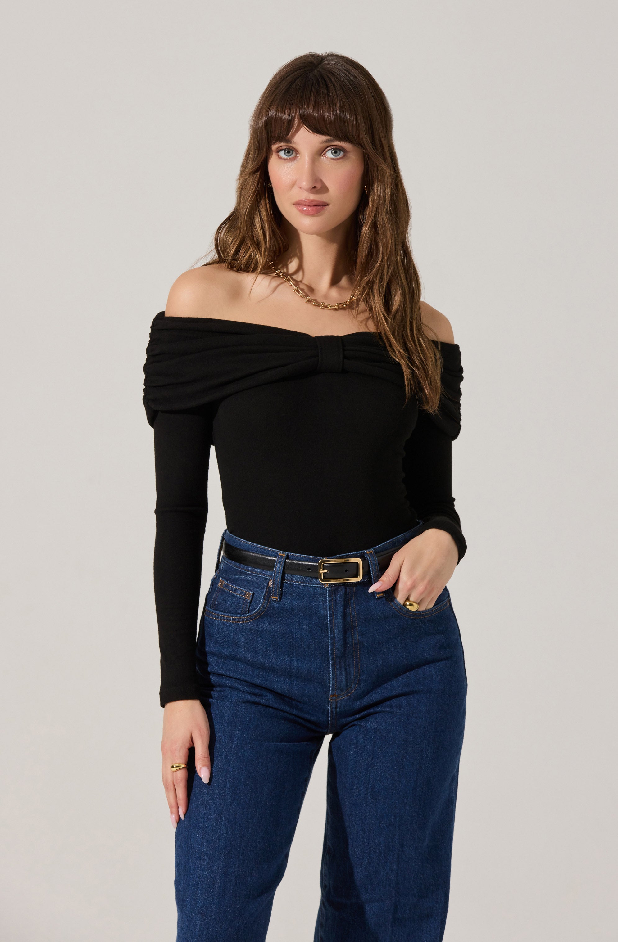 A woman is wearing a black off-the-shoulder top with long sleeves and a gathered, draped design across the shoulders. She pairs it with high-waisted, dark blue jeans that have a belt with a gold buckle. She accessorizes with a gold chain necklace and rings on her fingers.
