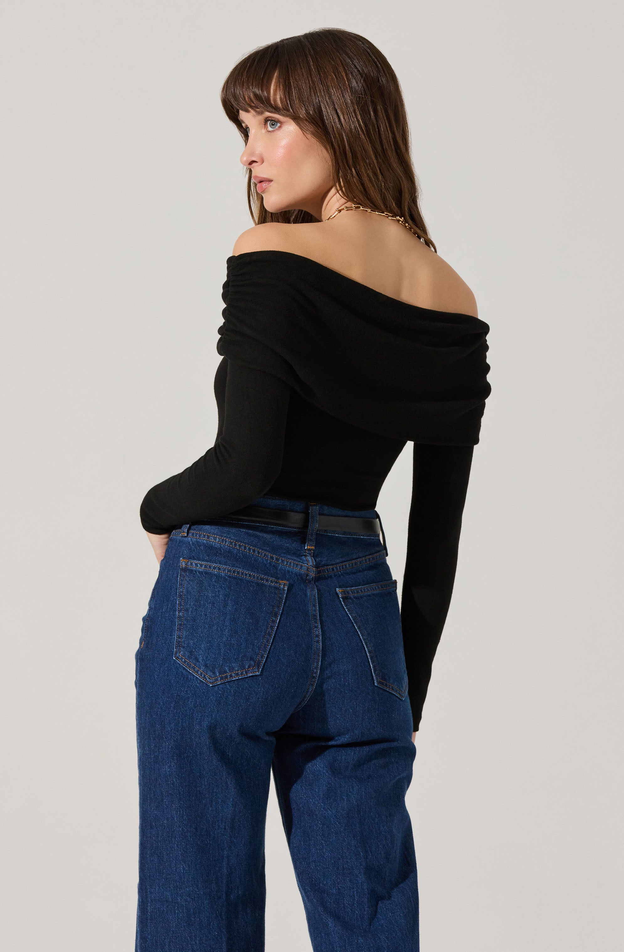 A woman is wearing an off-the-shoulder black top with long sleeves that are fitted, and the fabric appears to be soft and slightly stretchy. The top has a draped neckline that sits off her shoulders, exposing her collarbones. She pairs the top with high-waisted blue jeans that have a classic five-pocket style and a relaxed fit. The jeans sit comfortably at her waist, emphasizing her waistline.