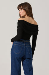 A woman is wearing an off-the-shoulder black top with long sleeves that are fitted, and the fabric appears to be soft and slightly stretchy. The top has a draped neckline that sits off her shoulders, exposing her collarbones. She pairs the top with high-waisted blue jeans that have a classic five-pocket style and a relaxed fit. The jeans sit comfortably at her waist, emphasizing her waistline.