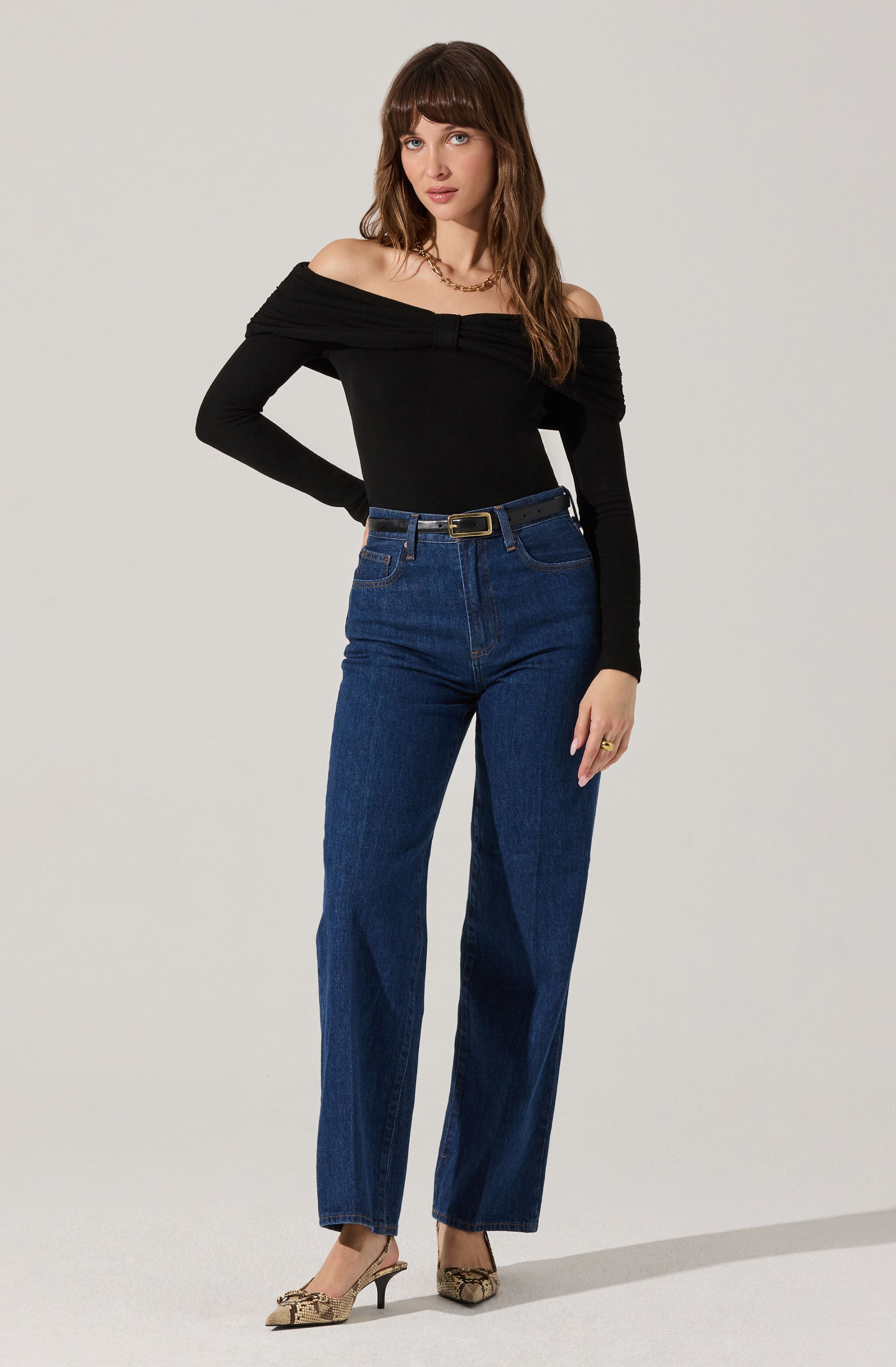 A woman is wearing a black, off-the-shoulder long-sleeve top with a gathered detail at the bust. She has paired it with high-waisted, straight-leg blue jeans that are secured with a black belt featuring a gold buckle. She is also wearing beige and black snakeskin-patterned pointed-toe high heels.