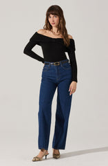 A woman is wearing a black, off-the-shoulder long-sleeve top with a gathered detail at the bust. She has paired it with high-waisted, straight-leg blue jeans that are secured with a black belt featuring a gold buckle. She is also wearing beige and black snakeskin-patterned pointed-toe high heels.