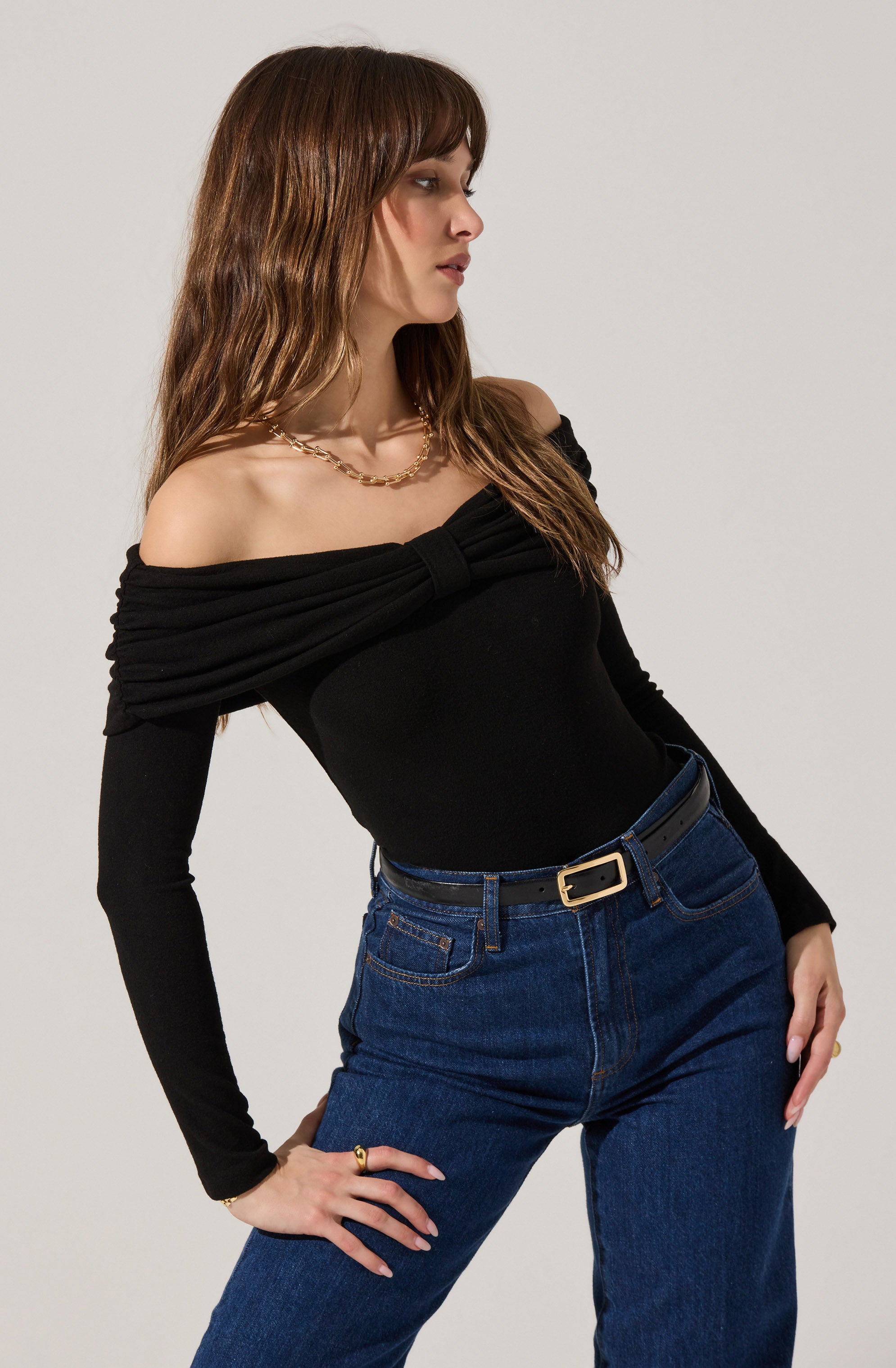 A woman is wearing an off-the-shoulder black top with long sleeves that are fitted and slightly ruched at the shoulders. She has paired it with high-waisted dark blue jeans and a black belt with a gold buckle. She is accessorized with a gold chain necklace.
