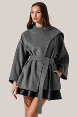 A woman is wearing a stylish, oversized gray coat with wide, long sleeves, a high neckline, and a matching belt tied around the waist. The coat features a layered design with different shades of gray and has a structured, modern silhouette. She pairs the coat with a short black skirt that is visible below the hem of the coat.