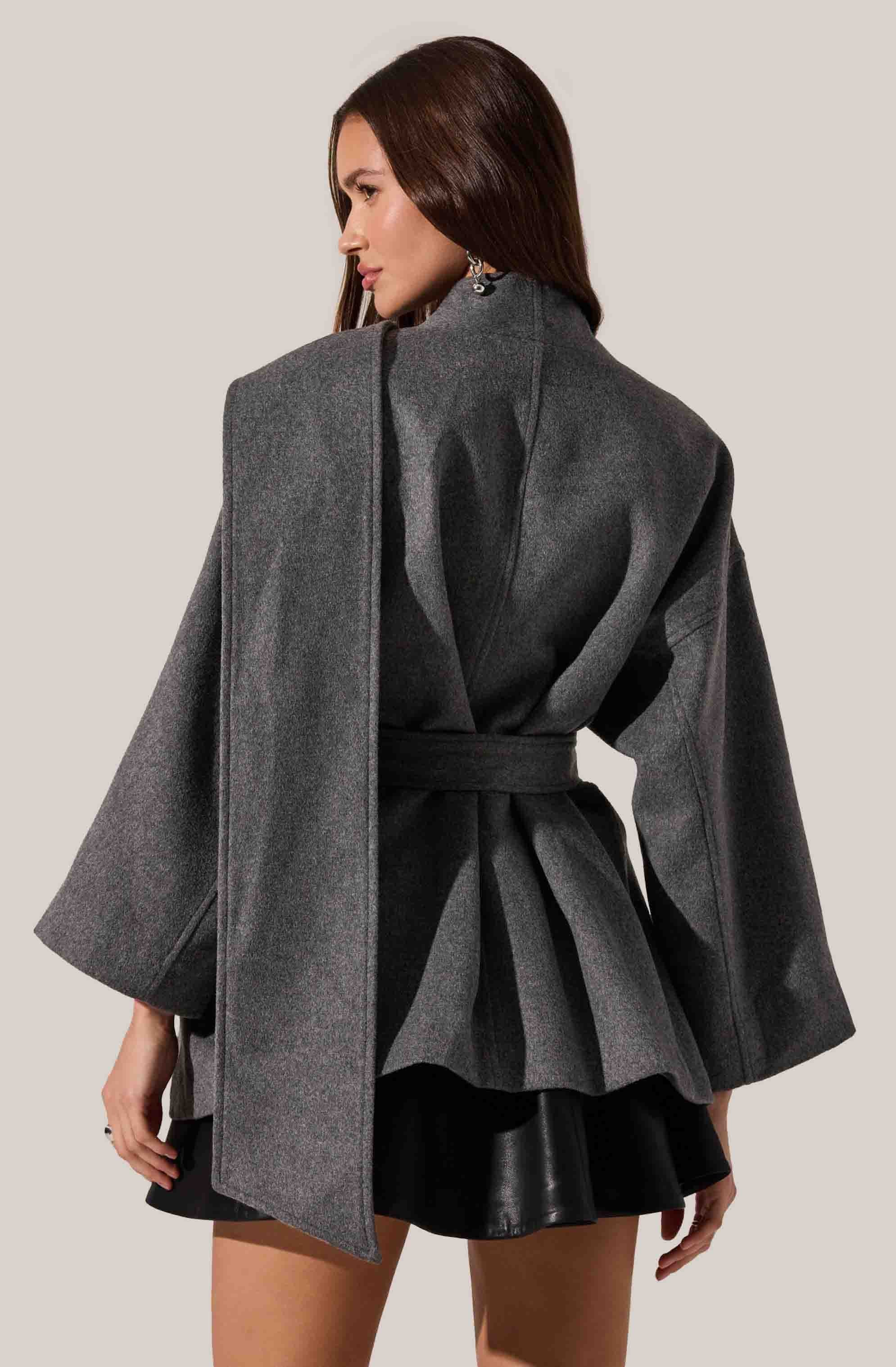 A woman is wearing a dark gray oversized coat with wide sleeves and a high collar, tied at the waist with a matching belt. Underneath, she has a black pleated skirt with a shiny finish. She is accessorized with dangling earrings.