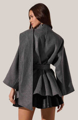 A woman is wearing a dark gray oversized coat with wide sleeves and a high collar, tied at the waist with a matching belt. Underneath, she has a black pleated skirt with a shiny finish. She is accessorized with dangling earrings.