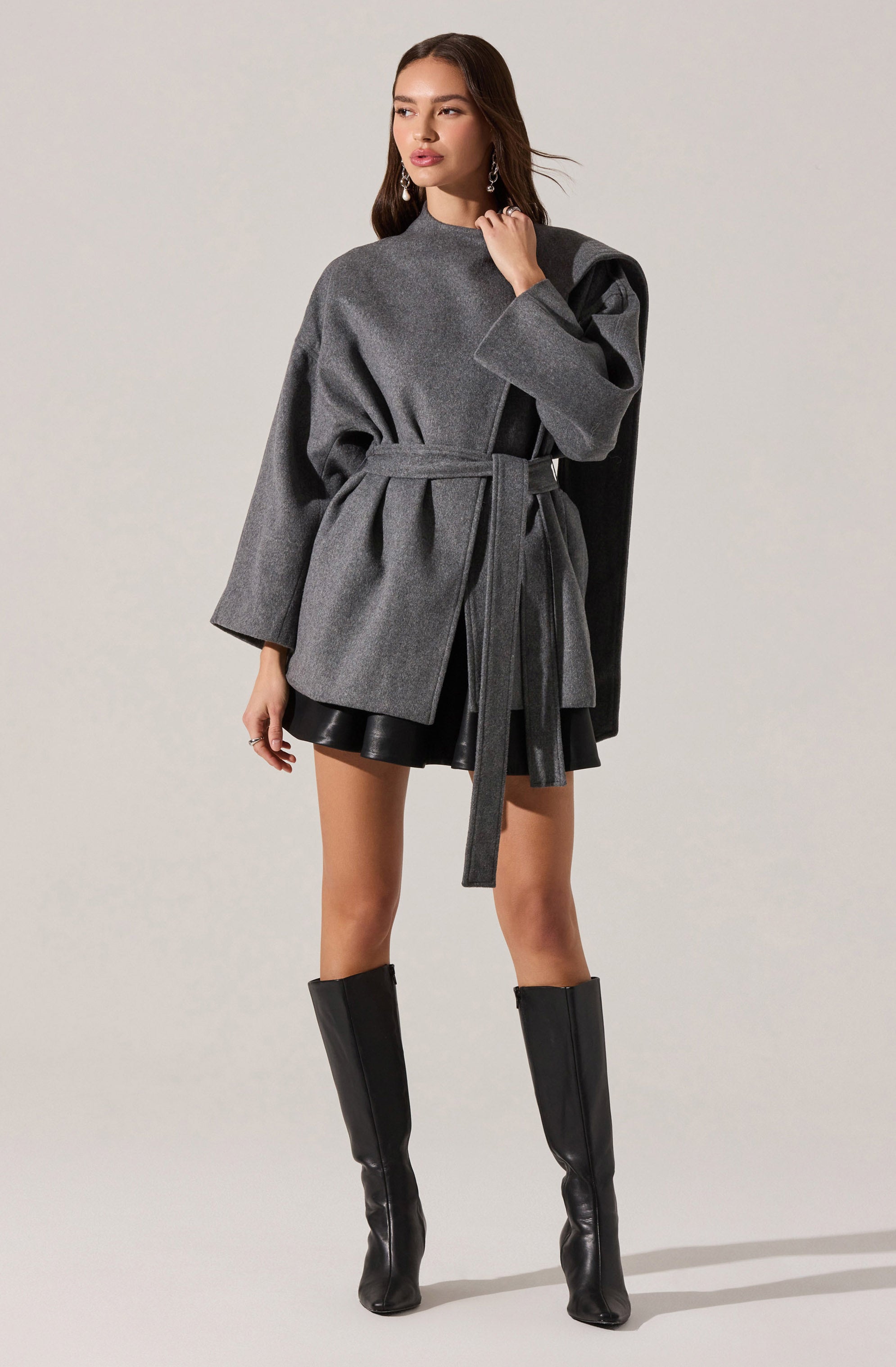 A woman is wearing a gray oversized coat with long sleeves and a belt at the waist. The coat has a darker gray panel on one shoulder, adding contrast. She pairs the coat with black leather shorts and knee-high black leather boots.