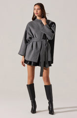 A woman is wearing a gray oversized coat with long sleeves and a belt at the waist. The coat has a darker gray panel on one shoulder, adding contrast. She pairs the coat with black leather shorts and knee-high black leather boots.