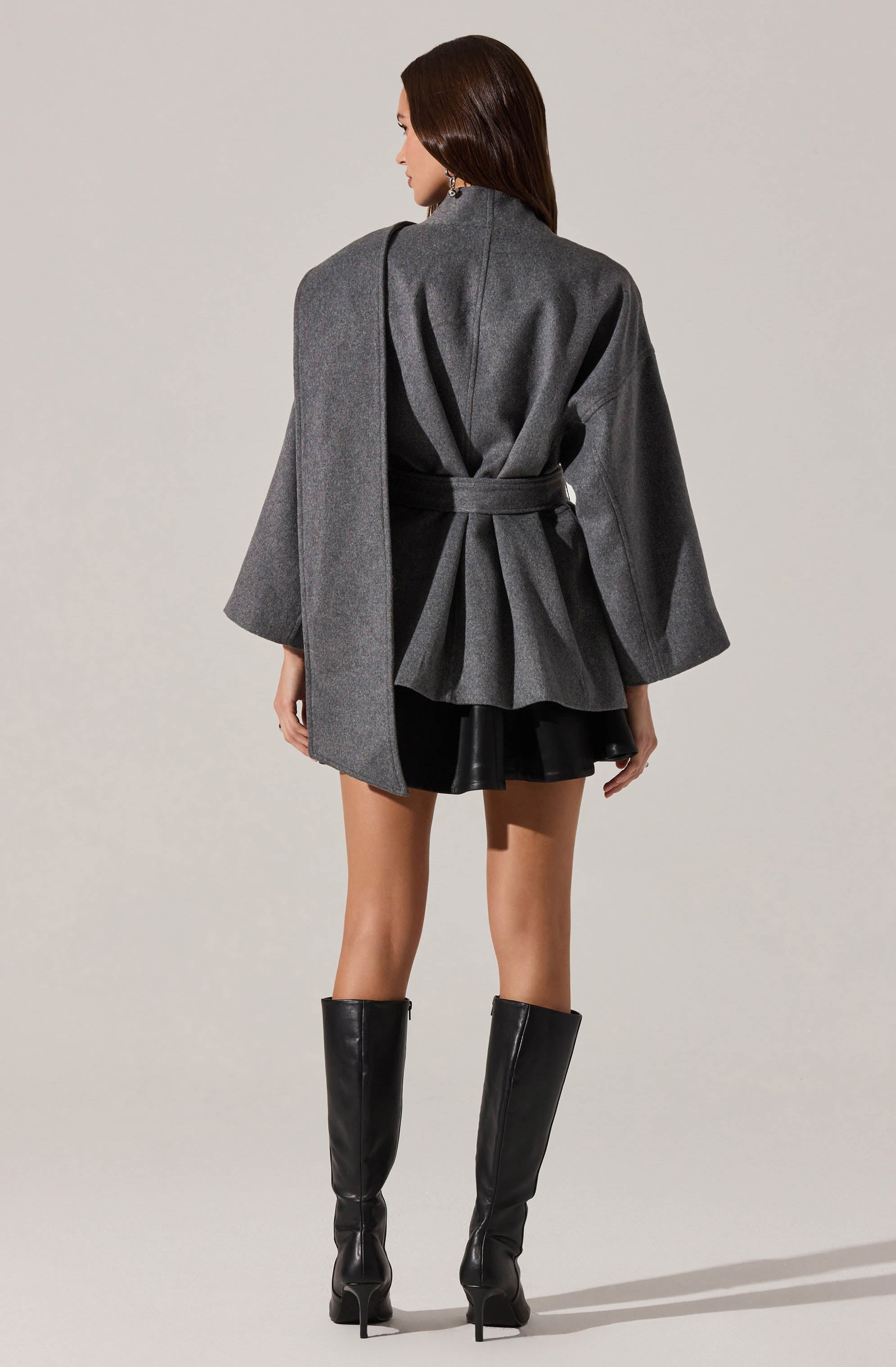 A woman is wearing a long, grey, wrap-style coat with wide sleeves that extend past her elbows, tied at the waist with a matching belt. She is also wearing a short black skirt and tall black leather boots with high heels.