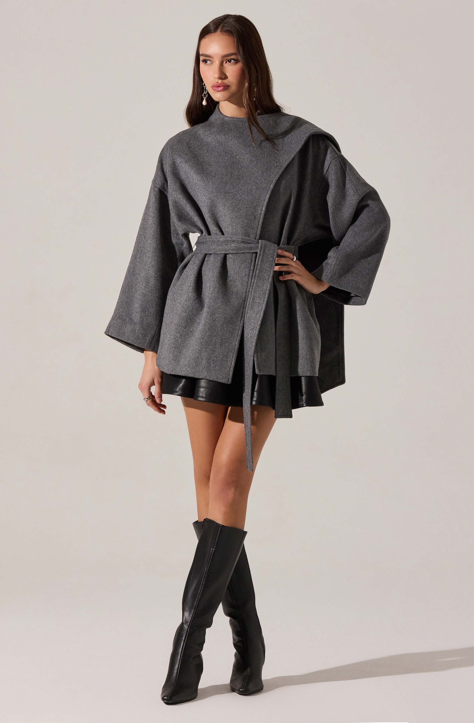 A woman is wearing a loose-fitting, gray, long-sleeved coat with wide sleeves and a high neckline. The coat is cinched at the waist with a matching fabric belt. She pairs it with a black mini skirt and knee-high black leather boots with zippers on the front.