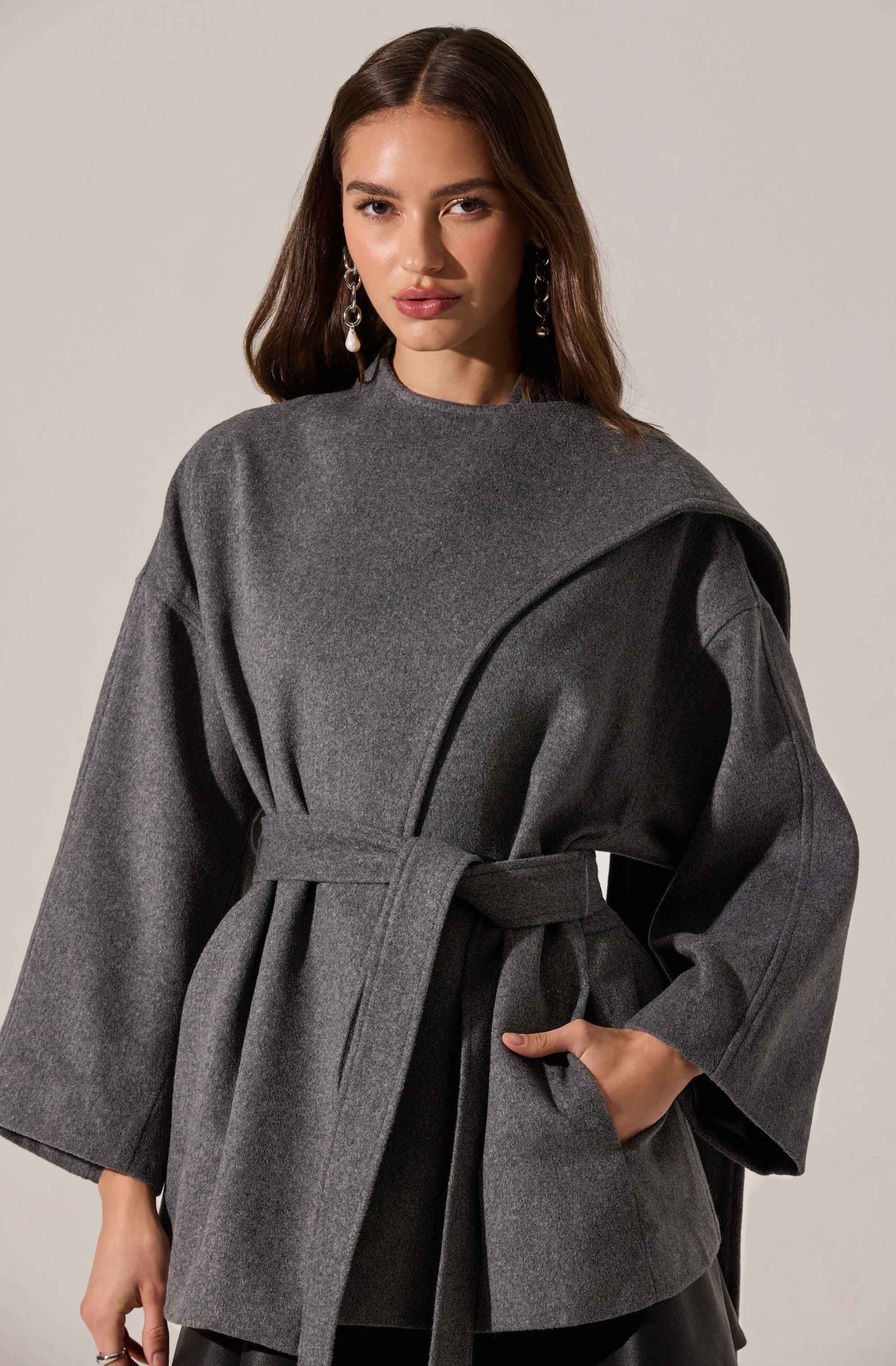 A woman is wearing a dark gray coat with wide sleeves and a waist tie. The coat features a high, rounded neckline and has a subtle drape detail on the shoulder. She is accessorized with long, dangling earrings.
