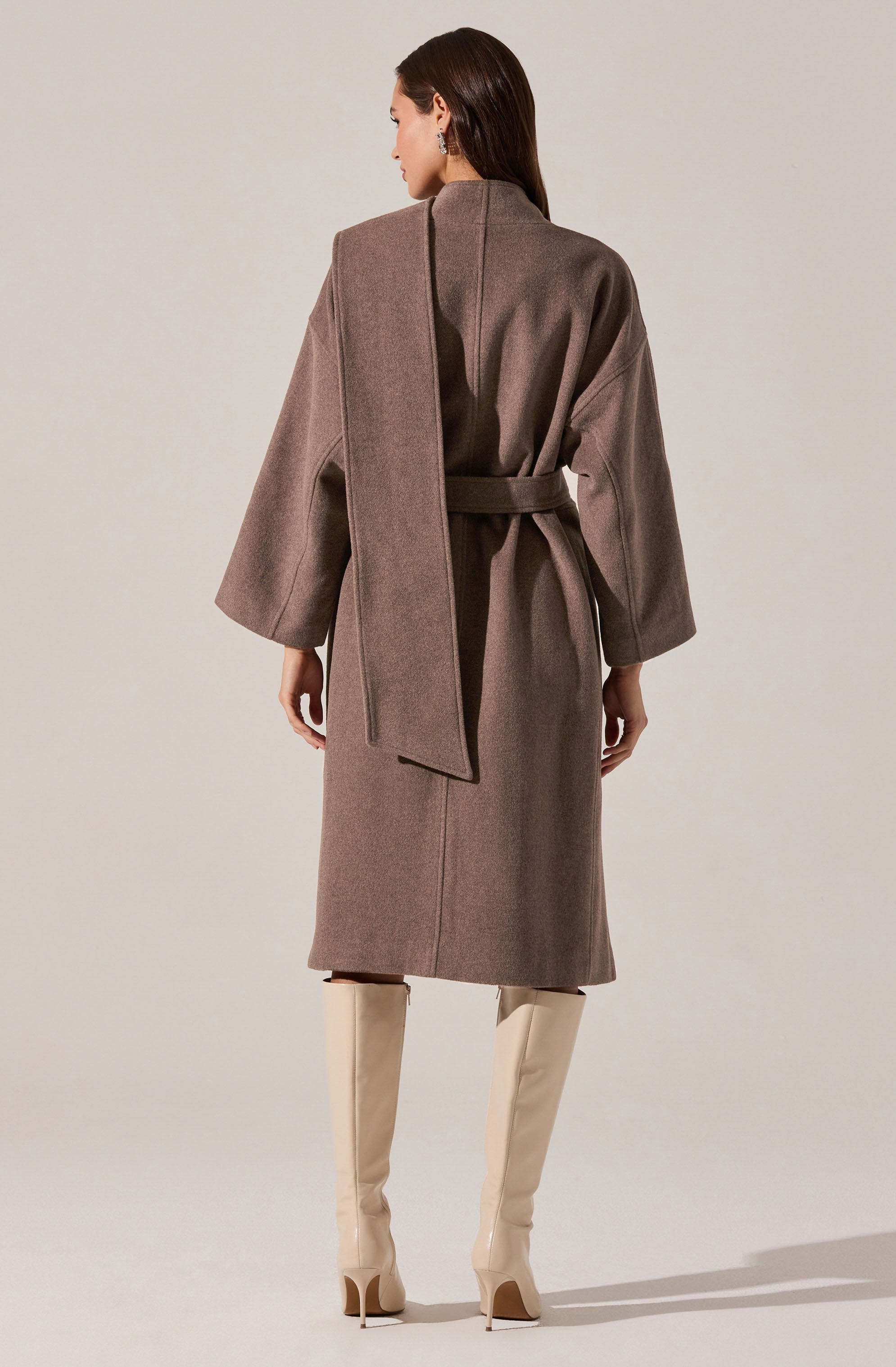 A woman is wearing a long, taupe-colored coat with wide sleeves and a matching belt tied at the waist. She has a scarf draped over her shoulders that matches the coat’s color. She is also wearing tall, cream-colored high-heeled boots.