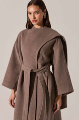 A woman is wearing a taupe-colored, oversized coat with wide sleeves. The coat features a large, draped collar and a matching fabric belt tied at the waist. The fabric appears to be a soft, wool or wool-blend material.