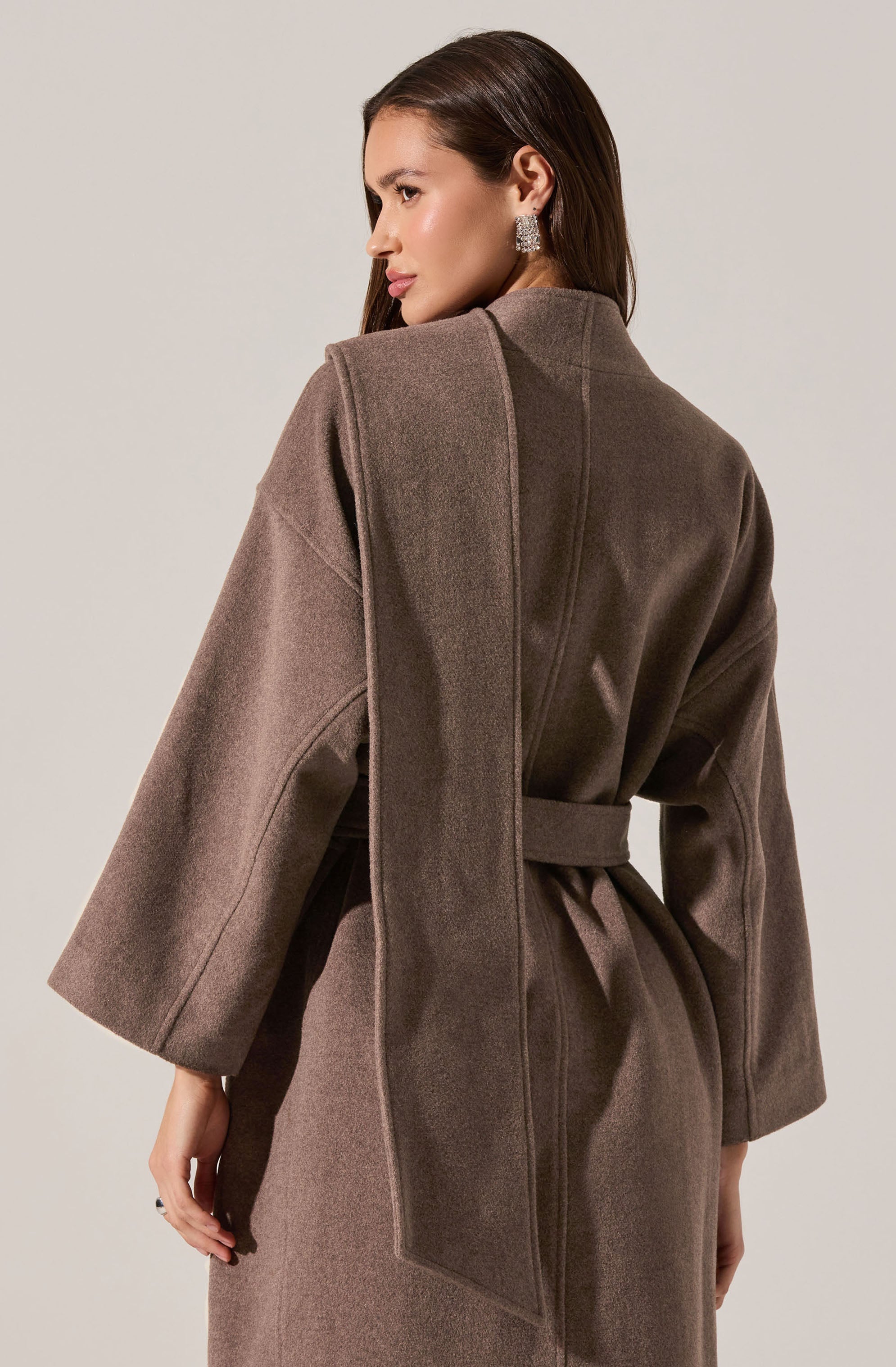 A woman is wearing a long, taupe-colored coat with wide, three-quarter length sleeves and a matching belt tied around the waist. The coat has a high collar and appears to be made of a heavy, warm fabric. She is accessorized with large, sparkly earrings.