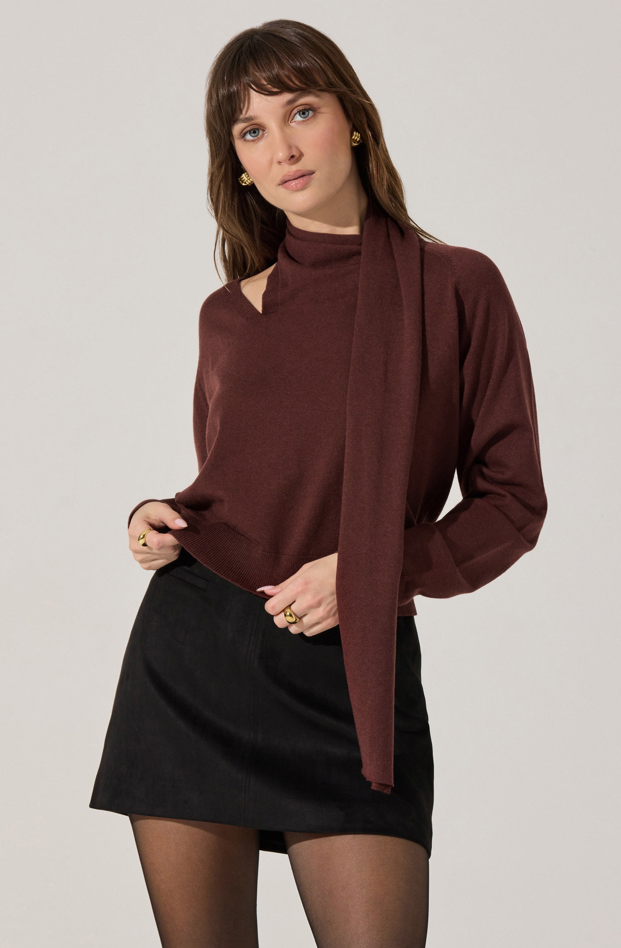 A woman is wearing a dark red, long-sleeved sweater with a matching scarf draped around her neck. She pairs it with a black mini skirt and sheer black tights. She accessorizes with gold earrings and rings.