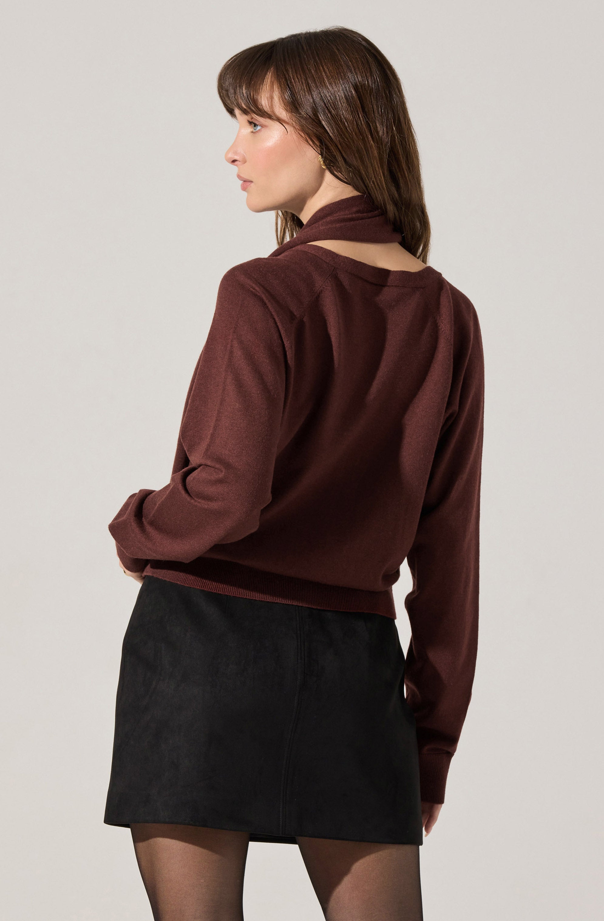 A woman is wearing a long-sleeved, burgundy sweater with a high neckline and a cutout detail at the back. She has paired it with a black mini skirt and sheer black tights.
