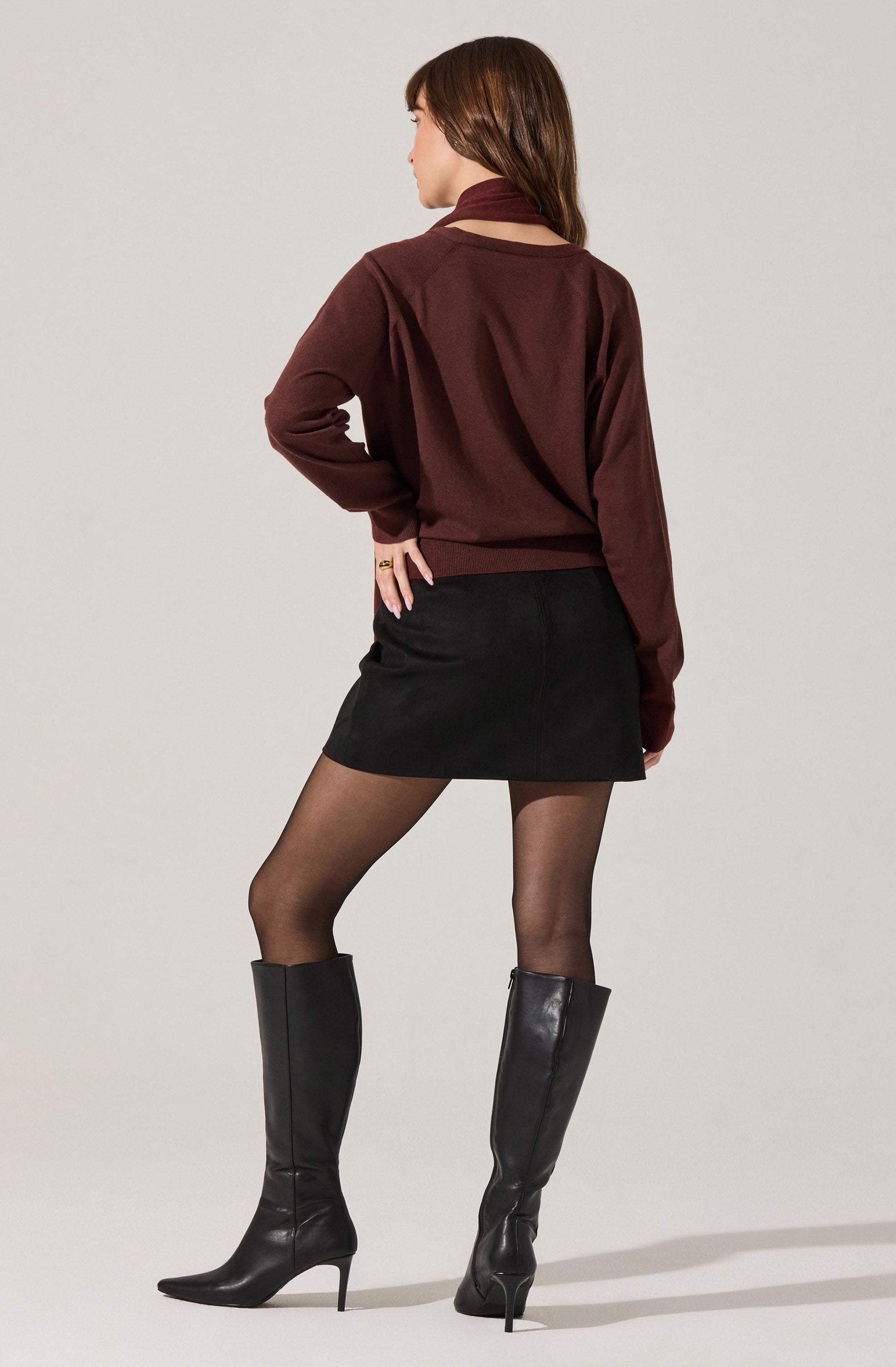 A woman is wearing a long-sleeve, burgundy sweater with a matching neck scarf, a black mini skirt, sheer black tights, and tall black leather high-heeled boots.