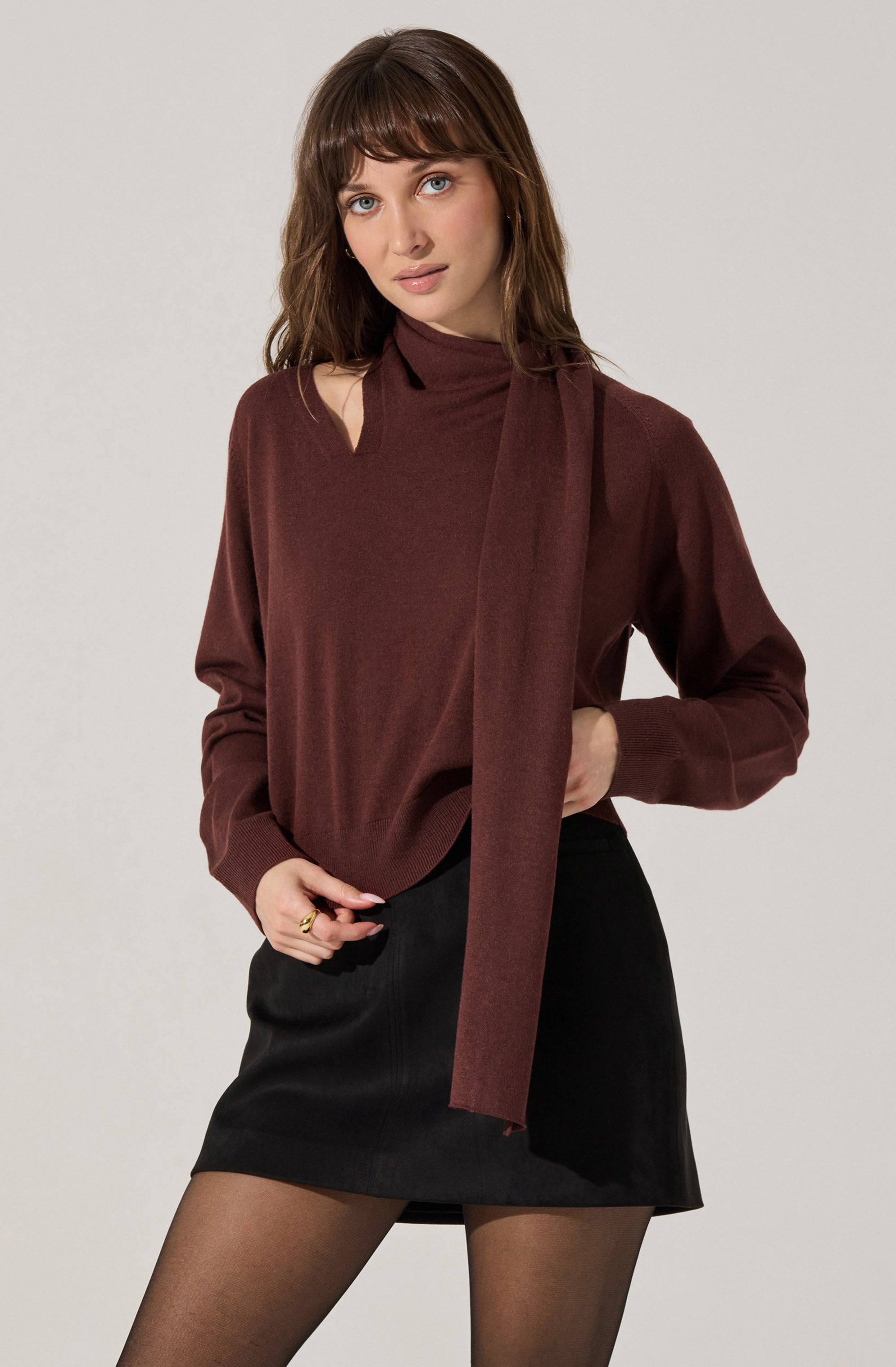A woman is wearing a long-sleeved, burgundy sweater with a matching scarf draped over her shoulder. She pairs it with a black mini skirt and sheer black tights.