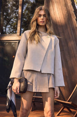 A woman is wearing a light beige, cropped blazer with long sleeves and a rounded collar. She has a small, gold brooch pinned on the left side of the blazer. She is also wearing matching beige, pleated shorts. She holds a black clutch bag with a gold chain strap in her left hand, which is accented with a patterned silk scarf tied around the wrist.
