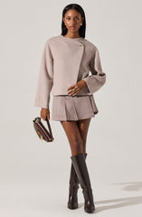 A woman is wearing a loose, long-sleeved, light beige top with a small decorative pin on the left side near the shoulder. She has paired it with a matching beige pleated mini skirt. She is also wearing black knee-high boots and is carrying a dark brown handbag with a gold chain strap and embellishments.