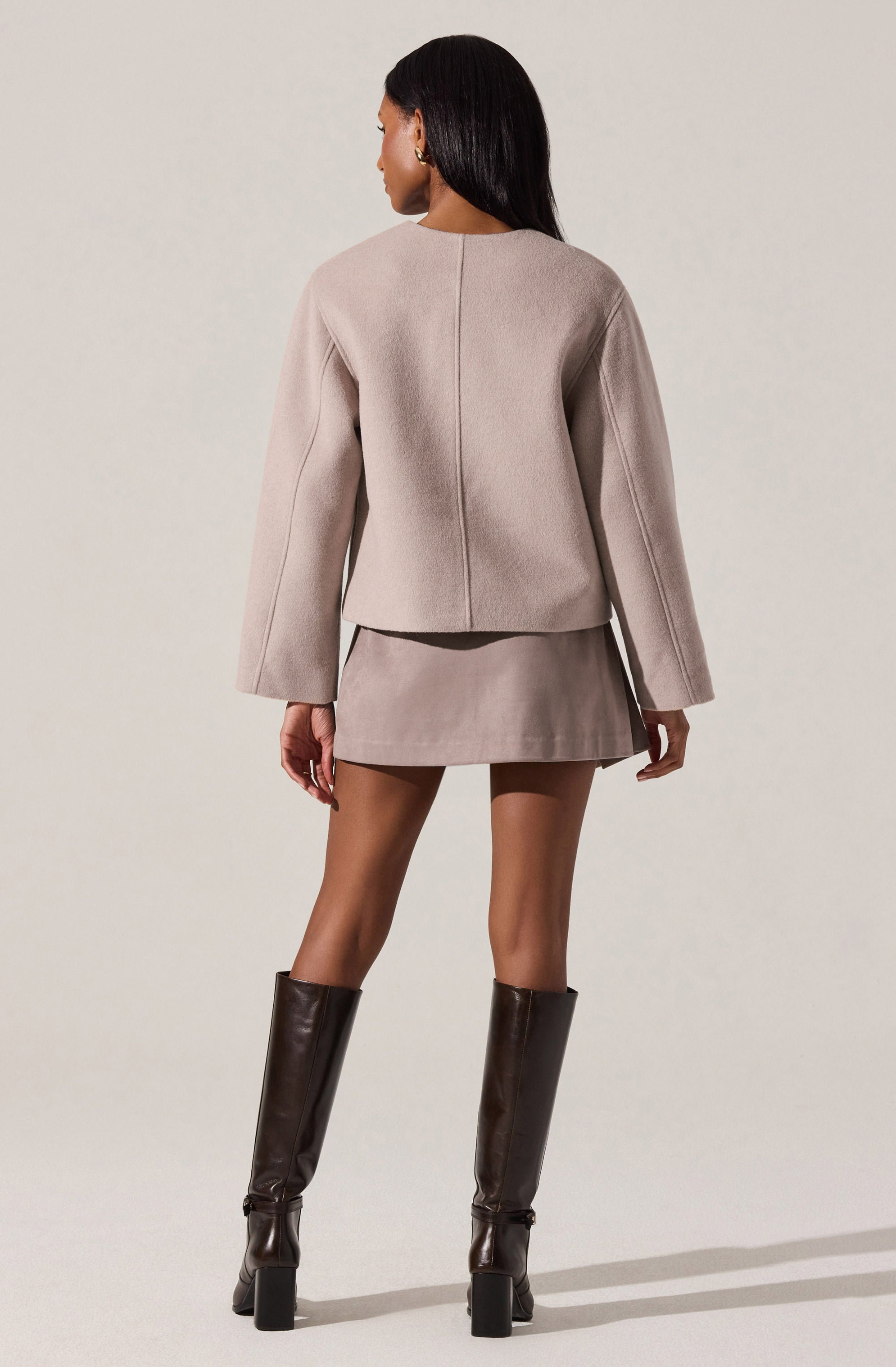 A woman is wearing a light beige, structured blazer with long, slightly flared sleeves and a subtle seam detail down the back. She pairs it with a matching mini skirt and tall, dark brown leather knee-high boots with block heels.