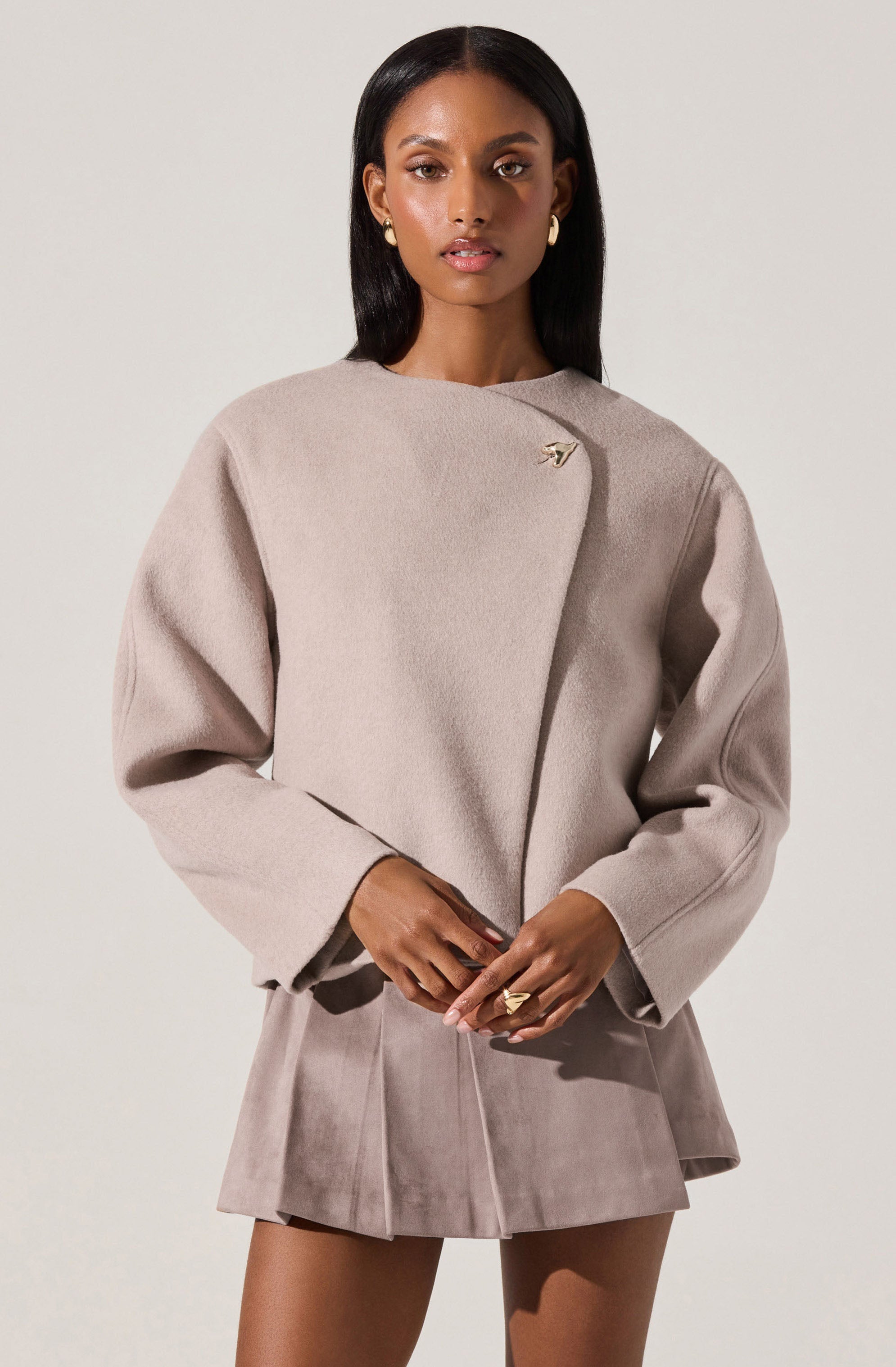 A woman is wearing a light beige, long-sleeved, structured coat with a high neckline and an asymmetrical closure secured by a decorative pin on the upper chest. She pairs it with a matching beige mini skirt with pleats, made from a soft, suede-like material. The outfit is complemented by gold earrings and rings.