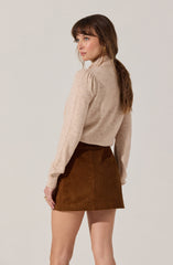 A woman is wearing a beige sweater with subtle embroidered details and puffed long sleeves, paired with a high-waisted, brown corduroy A-line mini skirt.
