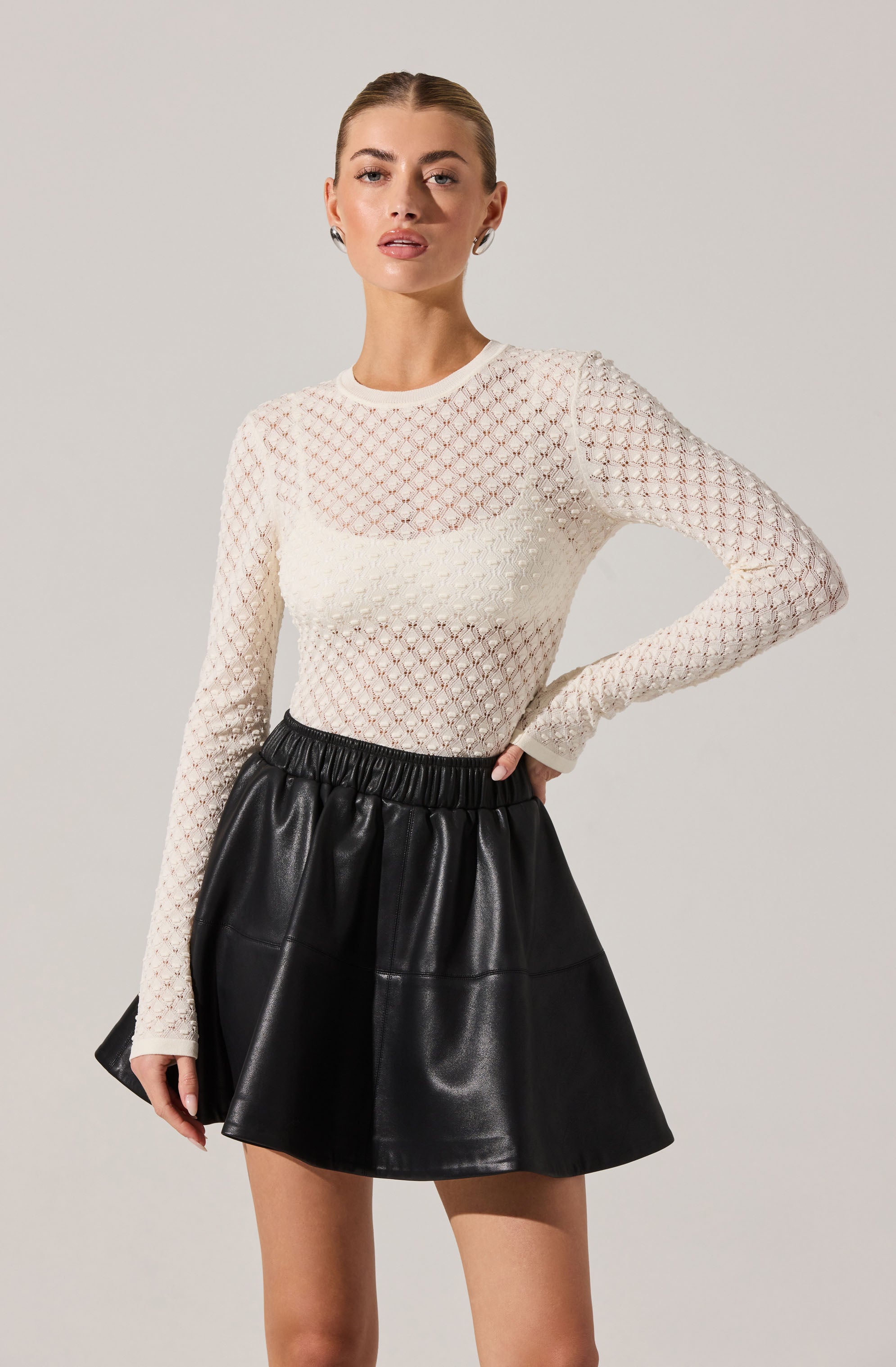 A woman is wearing a cream-colored, long-sleeved, open-knit sweater with a diamond pattern. The sweater is slightly sheer, revealing a layer underneath. She pairs it with a black, high-waisted, A-line mini skirt made of faux leather with a gathered waistband.