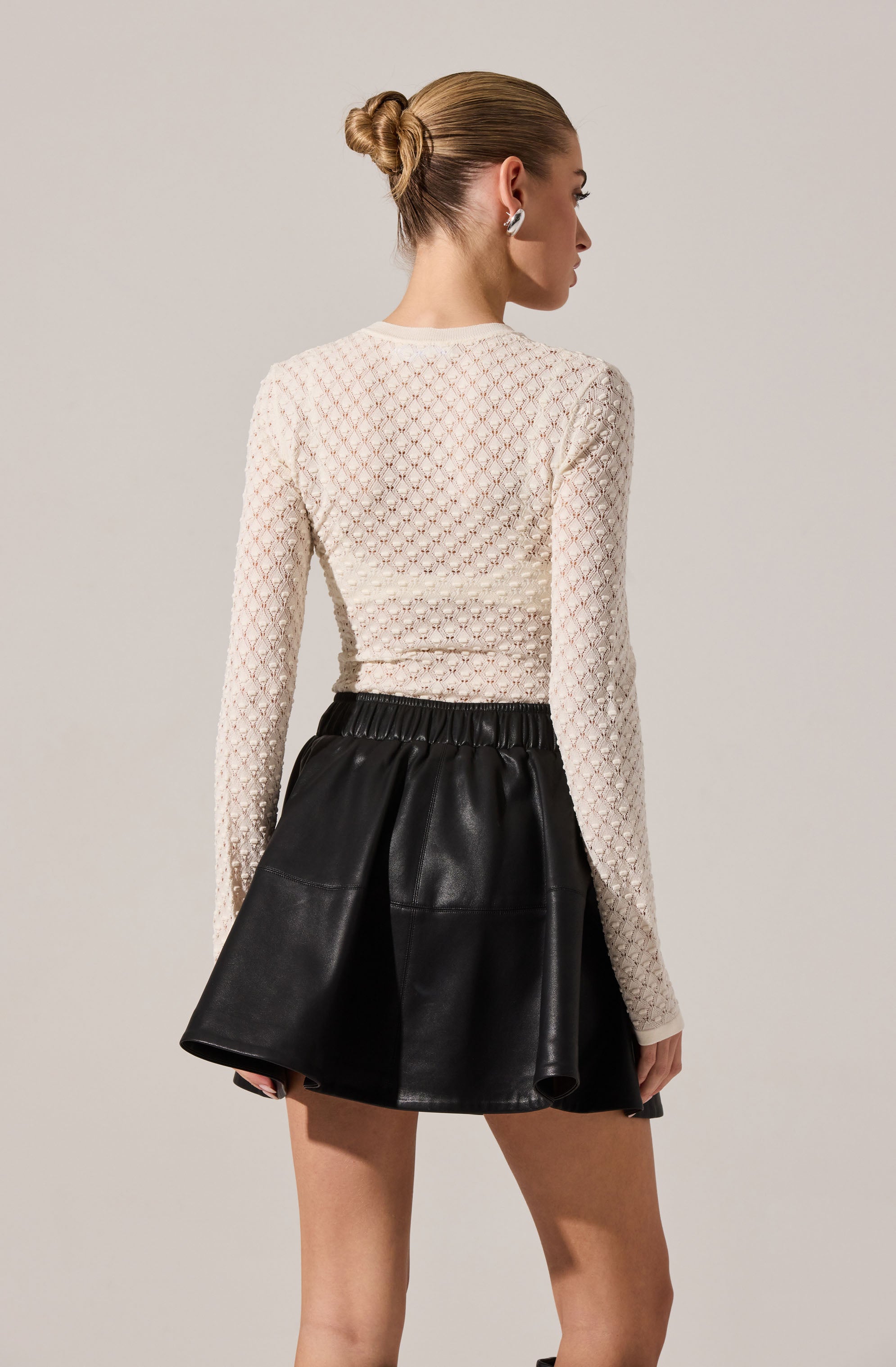 A woman is wearing a long-sleeved, white, textured knit top with a diamond lattice pattern. She pairs it with a high-waisted black leather skirt that has a gathered, flared design.