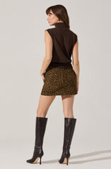 A woman is wearing a sleeveless, dark brown turtleneck vest paired with a short, leopard print skirt in shades of brown and black. She is also wearing tall, black, high-heeled boots that reach above her knees.
