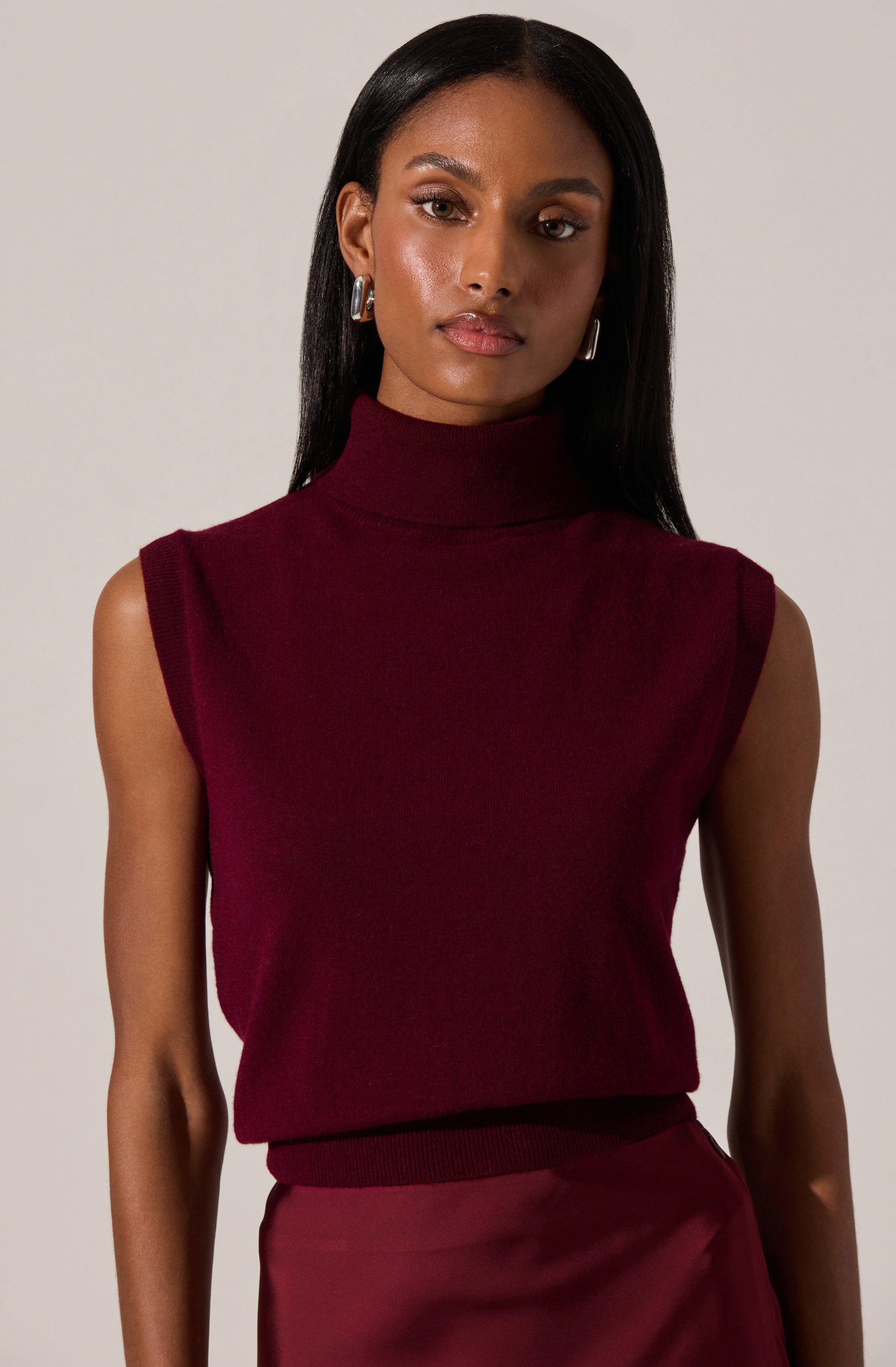 A woman is wearing a sleeveless, high-neck burgundy sweater with a ribbed hem and shoulder details. She pairs it with a matching burgundy satin skirt.