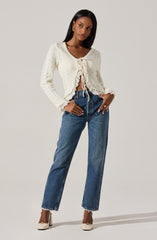 A woman is wearing an off-white, long-sleeved, knitted cardigan with a scalloped hem and ruffled cuffs, featuring a tie-front closure. She pairs it with high-waisted, straight-leg blue jeans and beige high-heeled shoes. She is also wearing gold earrings and a gold ring.