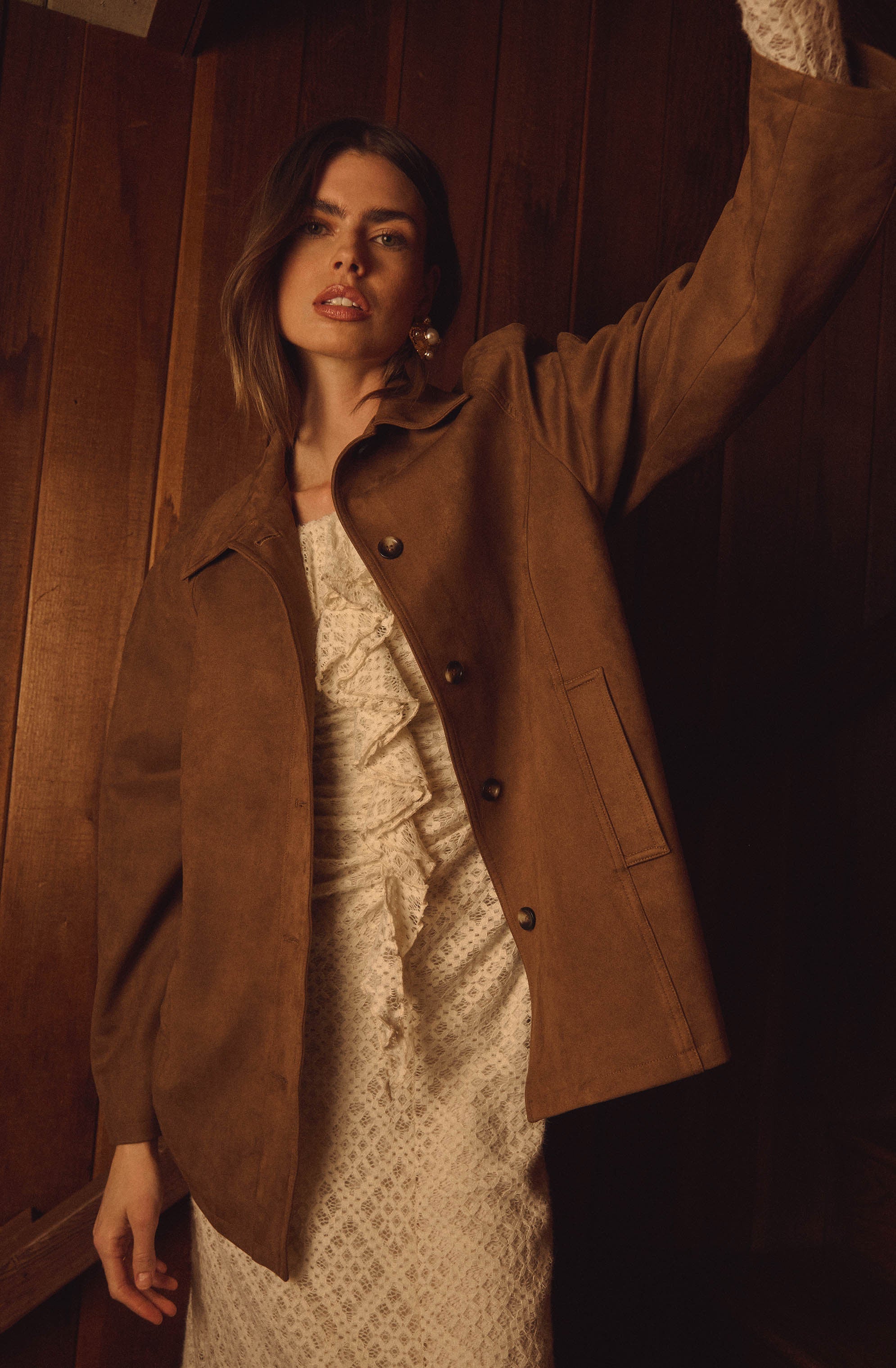 A woman wearing a brown suede jacket over a light-colored, patterned dress with a ruffled front. The jacket has four buttons and a large collar.