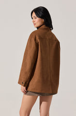A woman is wearing a loose-fitting, brown suede jacket with a collar and long sleeves. The jacket features buttoned cuffs and appears oversized. She is also wearing a short skirt with a small checkered pattern.