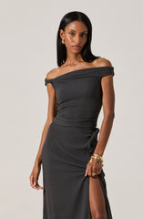 A woman is wearing an elegant off-the-shoulder black dress with a textured fabric. The dress has a draped neckline that sits off her shoulders and fits closely at the waist, flowing down to the floor with a high slit on one side. She accessorizes with gold earrings and multiple gold bracelets on her left wrist.