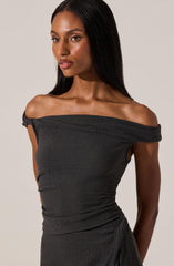 A woman is wearing a dark gray, off-the-shoulder dress made from a textured fabric. The dress features a wide neckline that drapes slightly over her shoulders and has a gathered, ruched appearance around the waist.