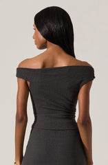 A woman is wearing a form-fitting, off-the-shoulder black dress with a textured fabric. The dress has a wide neckline that drapes over her shoulders, exposing her collarbones and upper back.