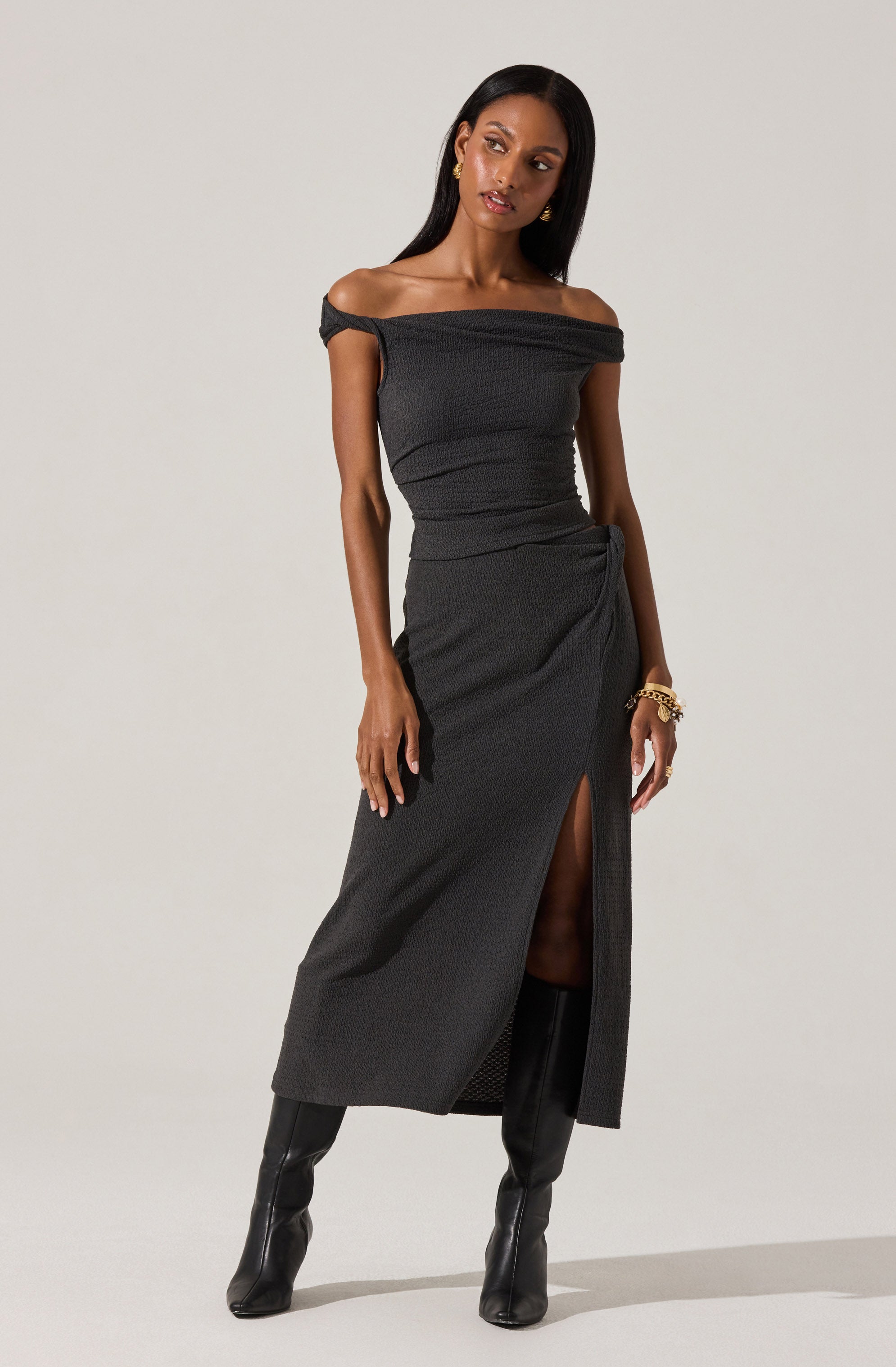 A woman is wearing an off-the-shoulder black dress with a textured fabric. The dress has a slit up the front, revealing one leg, and drapes slightly at the waist. She pairs the dress with tall black boots and accessorizes with gold jewelry, including earrings and bracelets.