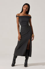 A woman is wearing an off-the-shoulder black dress with a textured fabric. The dress has a slit up the front, revealing one leg, and drapes slightly at the waist. She pairs the dress with tall black boots and accessorizes with gold jewelry, including earrings and bracelets.