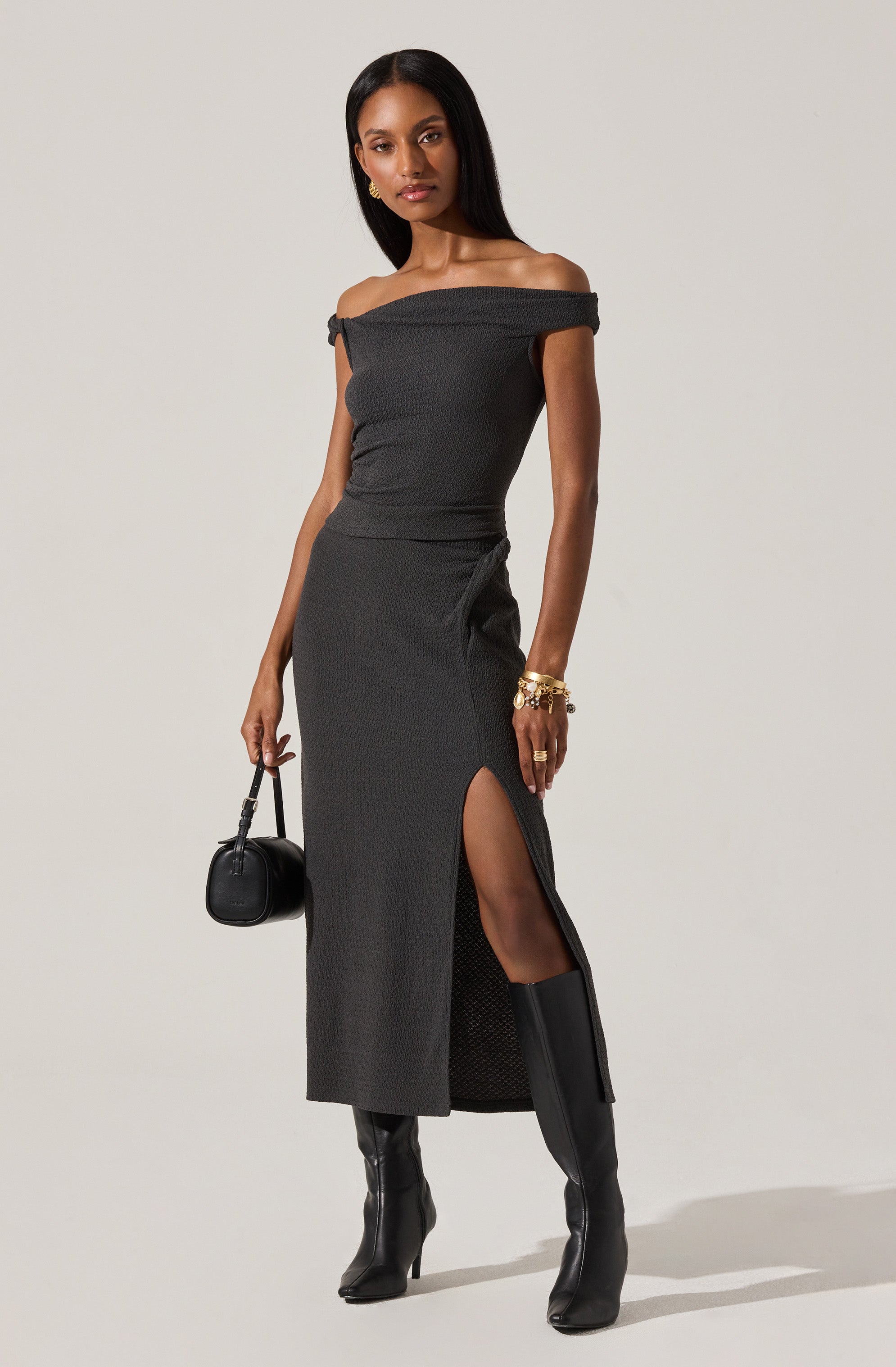 A woman is wearing an off-the-shoulder, short-sleeved, fitted black dress with a high slit on the side. She is accessorized with gold jewelry, including earrings and multiple bracelets. She is carrying a small black handbag and wearing knee-high black boots.
