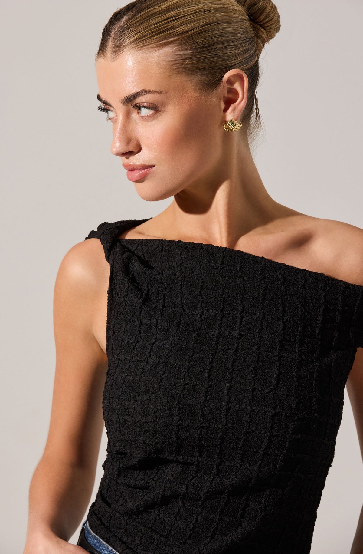 A woman is wearing a sleeveless black textured top with a wide, off-the-shoulder neckline. She has gold earrings, her hair is styled in a sleek bun, and she has a neutral makeup look.
