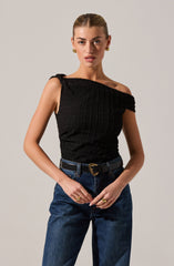 A woman is wearing a black sleeveless top with a textured, grid-like pattern and an asymmetrical, off-shoulder neckline. She pairs the top with high-waisted blue jeans and a black belt featuring a gold heart-shaped buckle. She accessorizes with gold earrings and multiple rings.