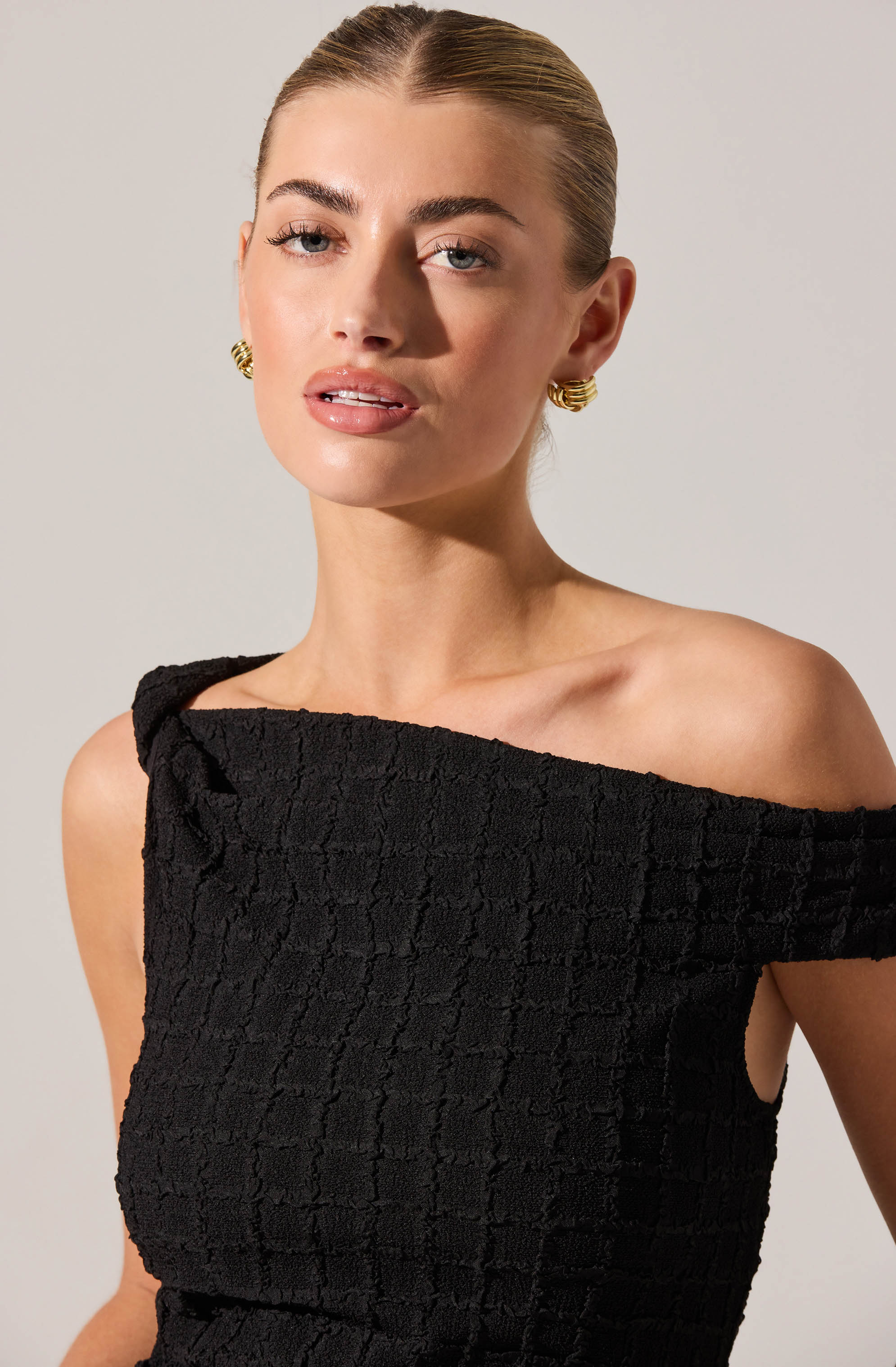 A woman with blonde hair styled in a sleek, pulled-back look is wearing a black, textured, off-the-shoulder dress. The dress has a square neckline and a unique shoulder strap on one side, revealing her collarbone and shoulder. She accessorizes with gold spiral earrings. The background is plain and light-colored.