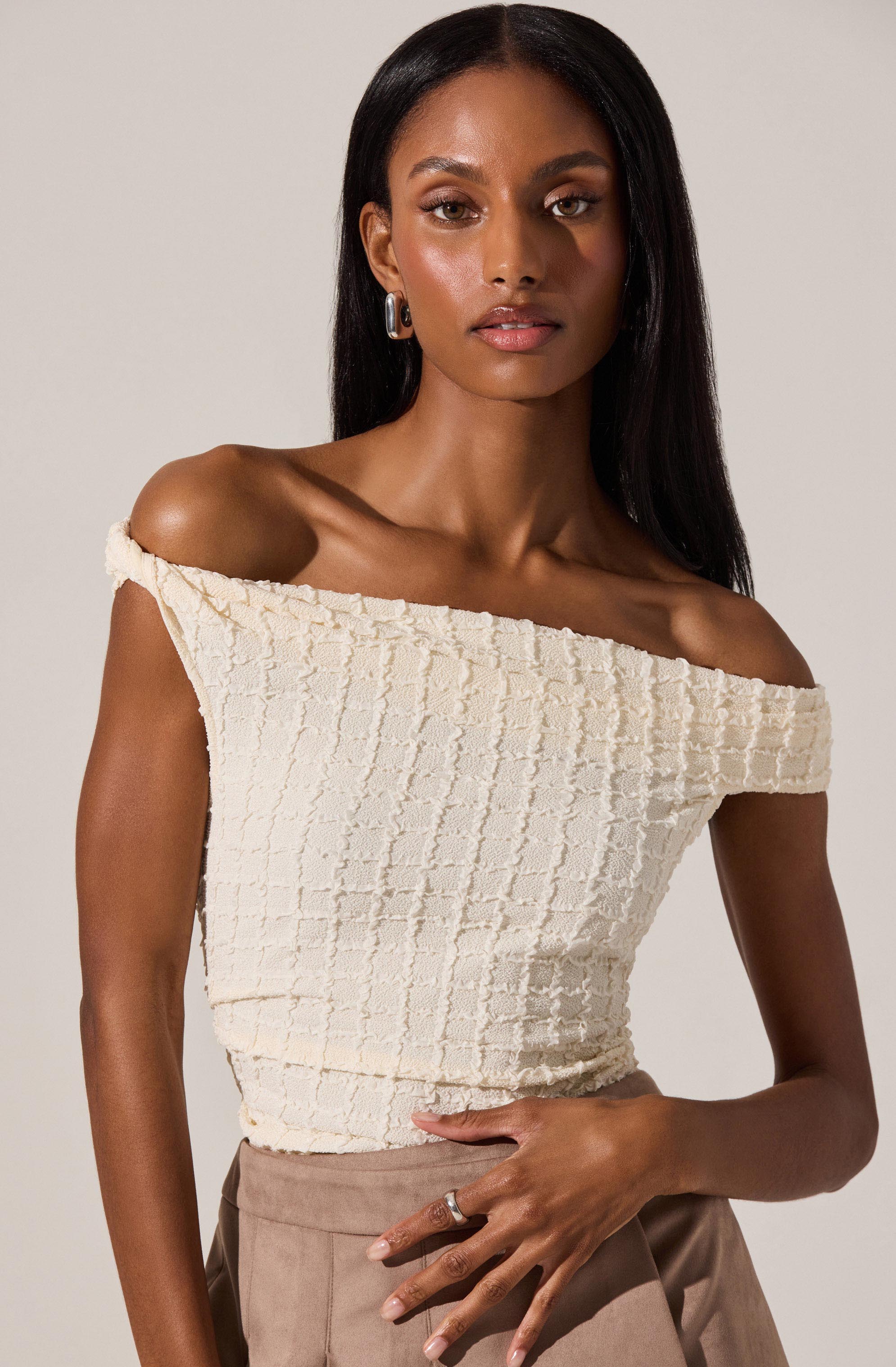 A woman is wearing an off-the-shoulder, cream-colored textured top with a grid pattern. The top has a relaxed fit, exposing her shoulders and collarbone. She pairs it with light brown high-waisted pants. She accessorizes with hoop earrings and a ring on her right hand.