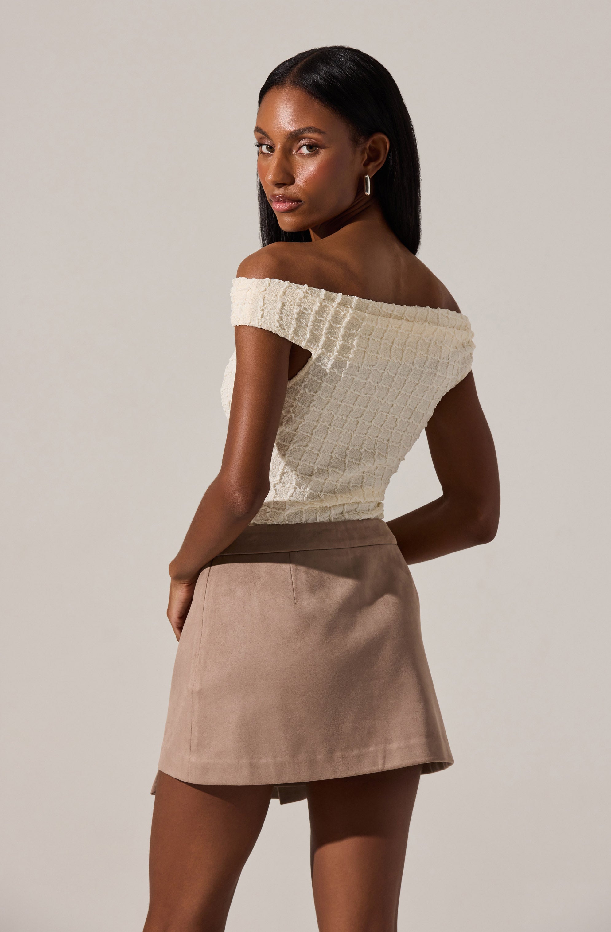 A woman is wearing an off-the-shoulder, short-sleeved white textured top with a grid pattern, paired with a high-waisted beige skirt that has a slight A-line shape.
