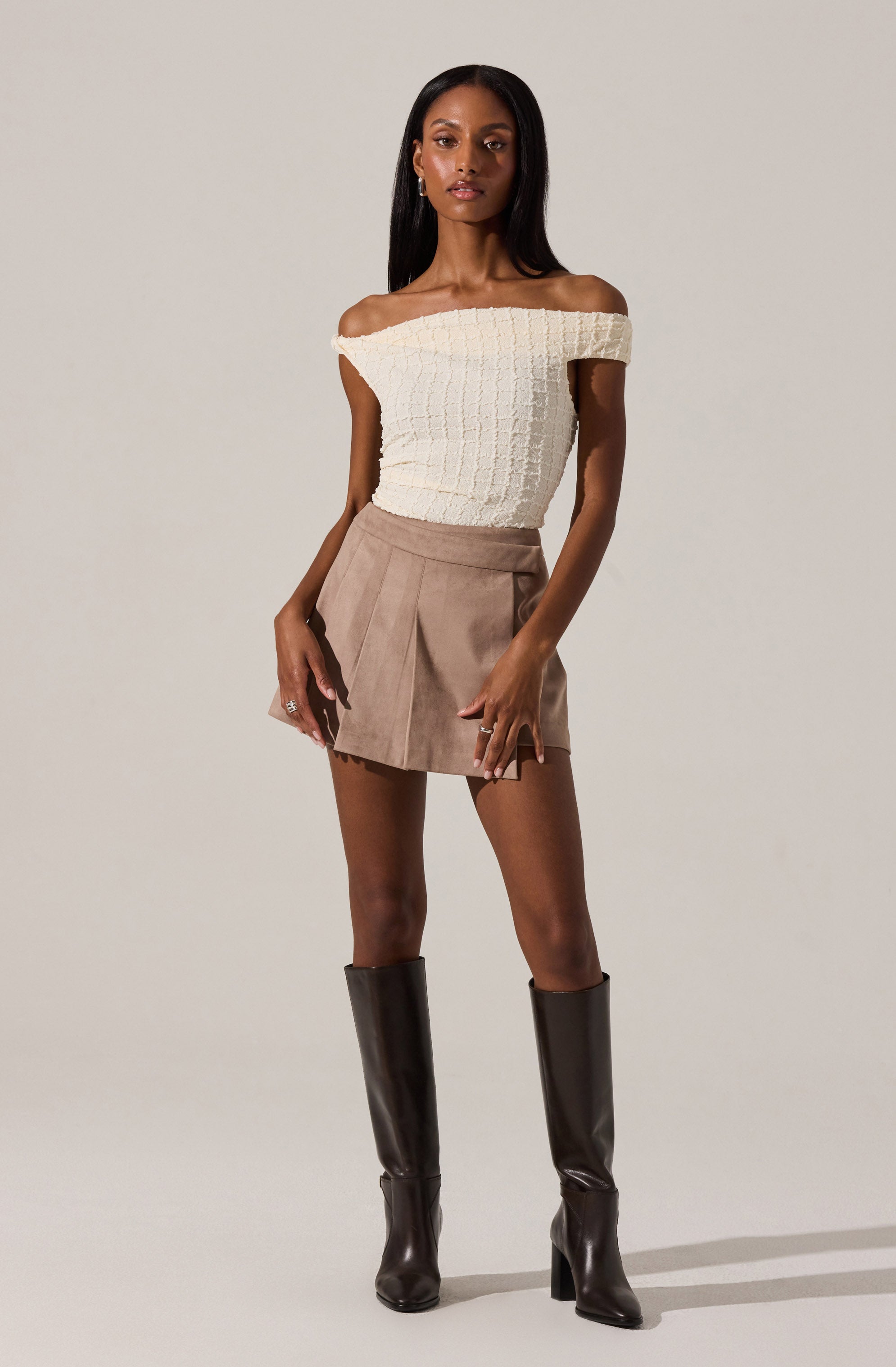 The woman is wearing a cream-colored off-the-shoulder knit top with a textured pattern, beige high-waisted shorts with pleats, and tall black leather knee-high boots with block heels.