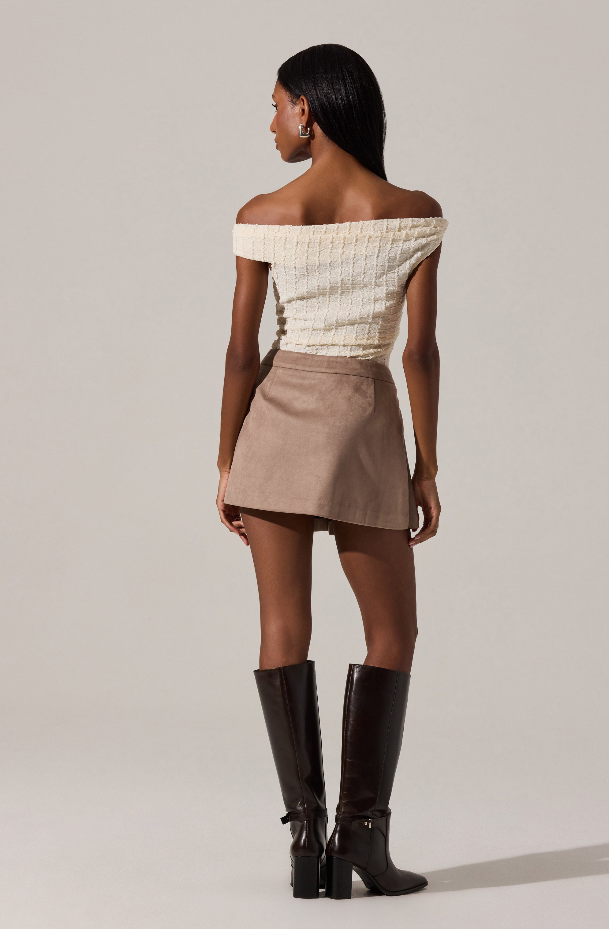 A woman is wearing an off-the-shoulder, cream-colored, textured top with a grid pattern. She pairs it with a taupe mini skirt and tall, dark brown leather boots with block heels.