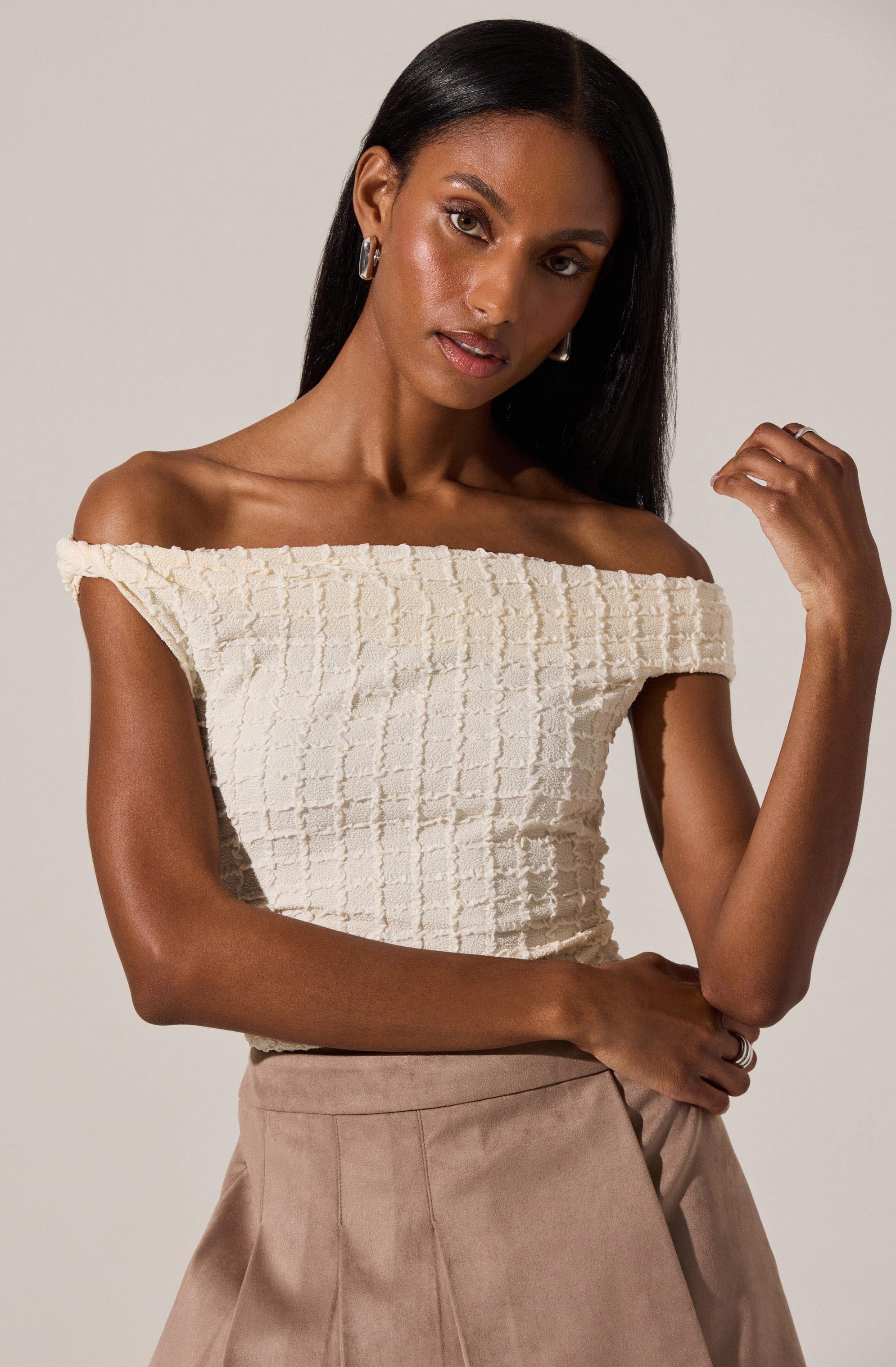 A woman is wearing an off-the-shoulder, cream-colored textured top with a grid-like pattern. She is also dressed in high-waisted, beige tailored pants.
