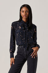 A woman is wearing a long-sleeved, dark-colored blouse with a celestial pattern that includes moons, stars, and clouds. The blouse features a bow tie at the collar. She has paired it with dark high-waisted jeans.