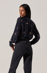 A woman is wearing a long-sleeved blouse with a dark background featuring a celestial print that includes moons, stars, and suns. She pairs it with high-waisted black jeans.
