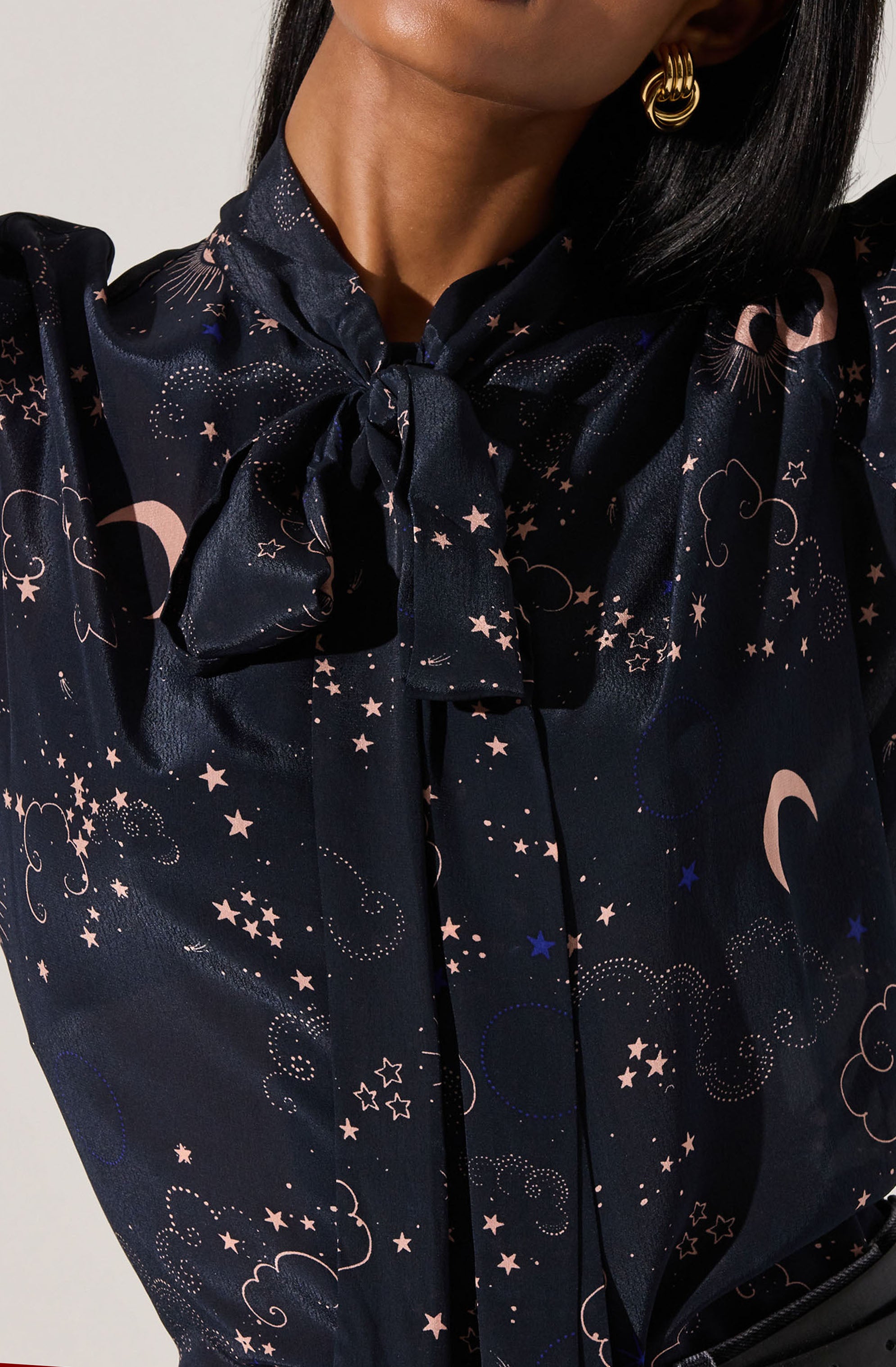 A woman is wearing a dark-colored blouse with a celestial pattern that includes moons, stars, and clouds in light pink, white, and blue. The blouse features a high neckline with a bow tied at the collar.